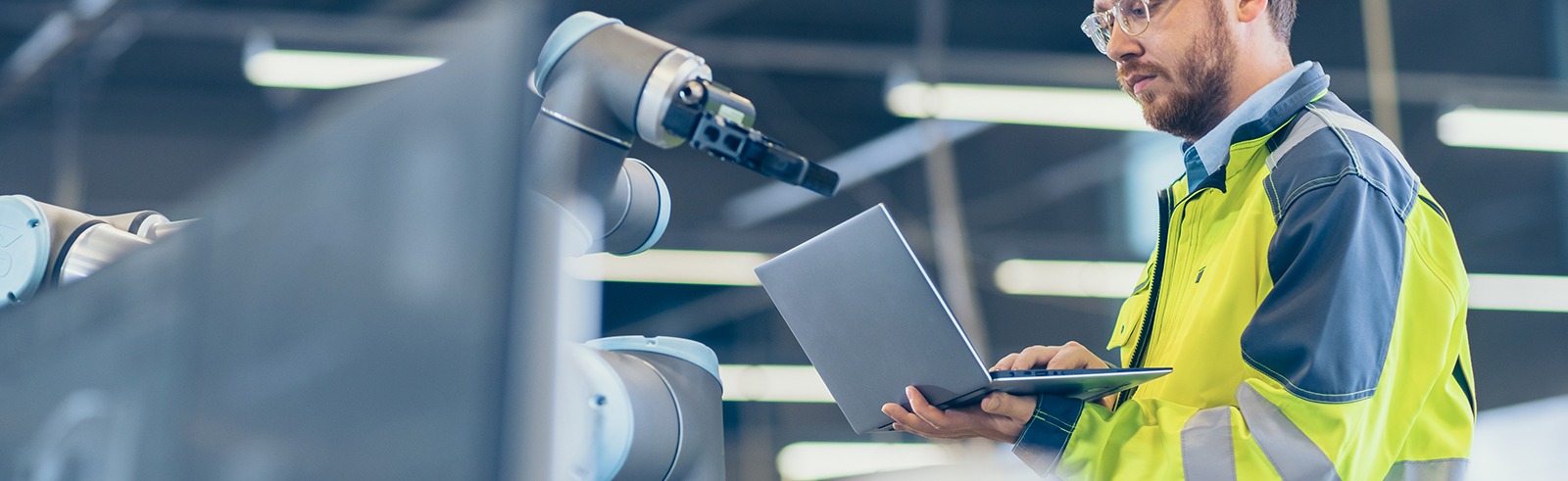 Technicien industriel en veste haute visibilité configurant un équipement à l’aide d’un ordinateur portable près d’un bras robotisé