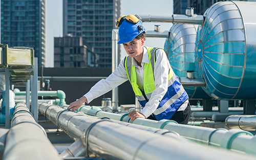 Technicien équipé de casque et gilet haute visibilité inspectant des conduites vapeur isolées sur une installation industrielle