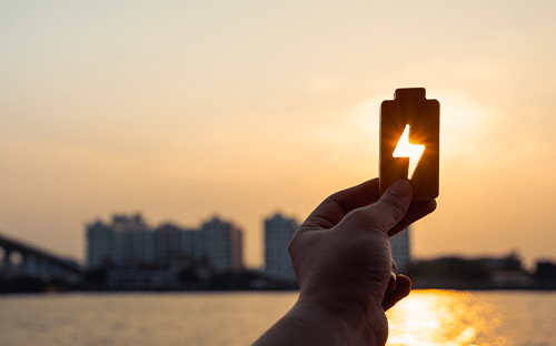 Mano che regge un'icona raffigurante una batteria con il simbolo di un fulmine davanti a un paesaggio urbano al tramonto, a simboleggiare il risparmio energetico.