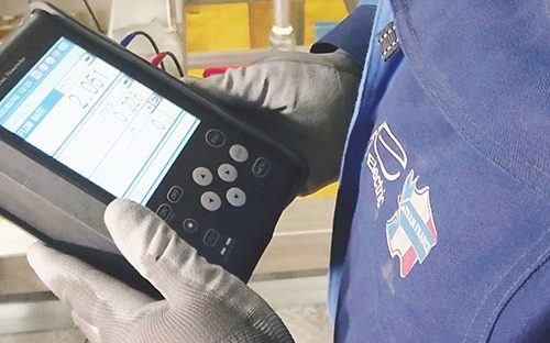 Technician holding a portable diagnostic device displaying measurements for flow meter inspection and maintenance