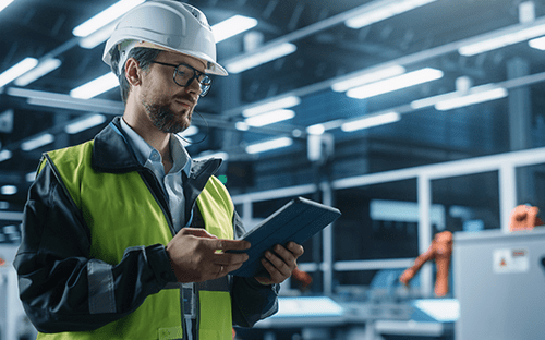 Industrietechniker mit Schutzhelm und Sicherheitsweste, der in einer automatisierten Werkstatt auf einem Tablet Energieverbrauchsdaten abruft