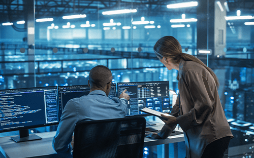 Two engineers analyze data on screens in front of a server room in the background.