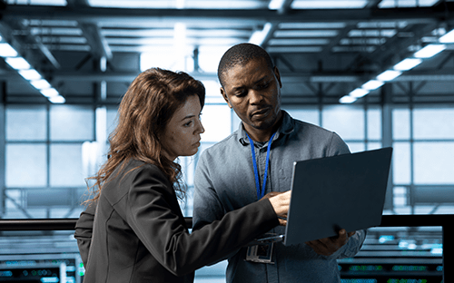Two technicians analyze data on a laptop in the server room.
