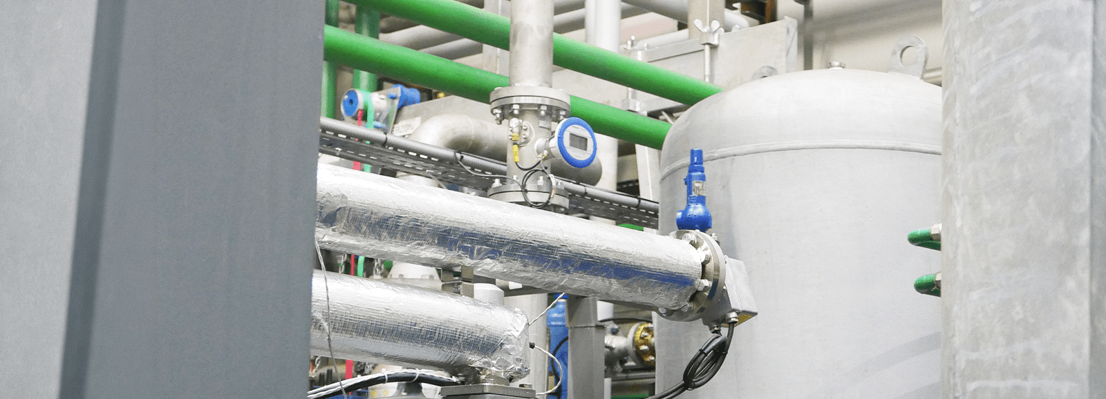 Ultraschall-Durchflussmesser, installiert an einer isolierten Rohrleitung in einer industriellen Umgebung mit Behälter und Rohrleitungsnetz