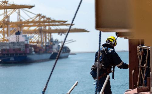 Techniker, der mit einem Sicherheitsgurt ausgestattet ist und an einer Schiffsstruktur in der Nähe von Hafenkränen Wartungsarbeiten an der Hydraulik durchführt