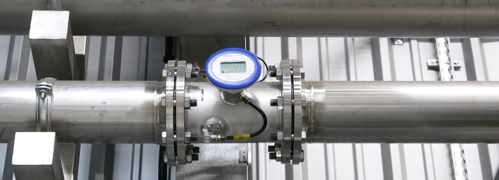 Medidor de caudal montado numa tubagem de aço inoxidável com visor digital, instalado entre duas flanges num ambiente industrial