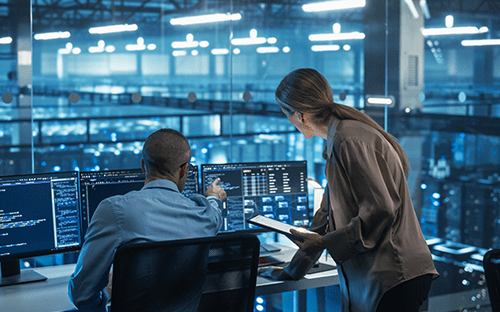 Arka planda sunucuların görüldüğü bir veri merkezinde, iki teknisyen izleme ekranlarını inceliyor