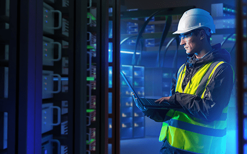 Bir sunucu odasında dizüstü bilgisayar kullanan, kask ve güvenlik yeleği giymiş bir teknisyen