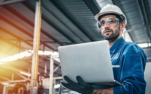 Ein Ingenieur mit Schutzhelm und Schutzbrille, der in einer industriellen Umgebung einen Laptop bedient