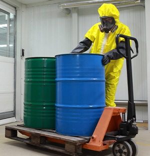 Arbeiter in einem Chemikalienschutzanzug, der mit einem Hubwagen zwei Metallfässer auf einer Palette bewegt (1)