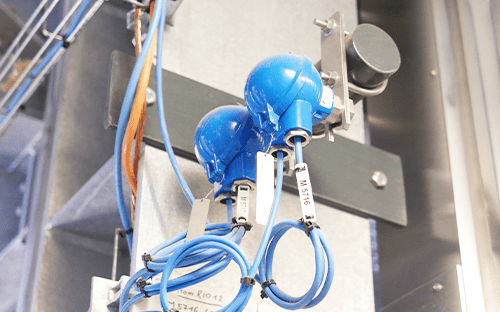 Zwei industrielle Temperaturfühler mit blauem Anschlusskopf, montiert in einer Anlage mit Verkabelung und Instrumentierung