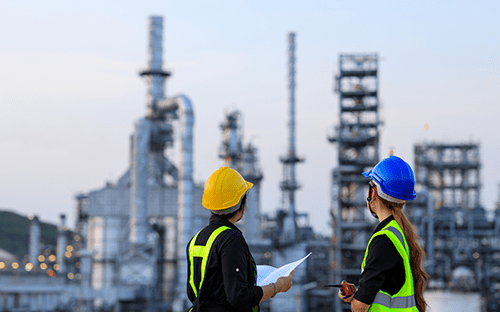 Deux techniciens en &eacute;quipements de protection observant une installation industrielle avec plans de contr&ocirc;le devant un site de production