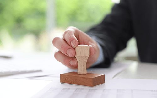 Eine Hand, die einen Holzstempel auf Verwaltungsunterlagen drückt, die auf einem Schreibtisch liegen