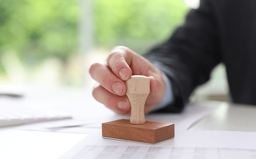 Eine Hand, die einen Holzstempel auf ein Dokument drückt, das auf einem Schreibtisch in einer Büroumgebung liegt