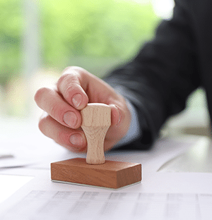 Main d’un professionnel apposant un tampon sur un document administratif posé sur un bureau
